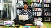 A woman sitting on the floor with her laptop and with books around her and a library shelf behind.