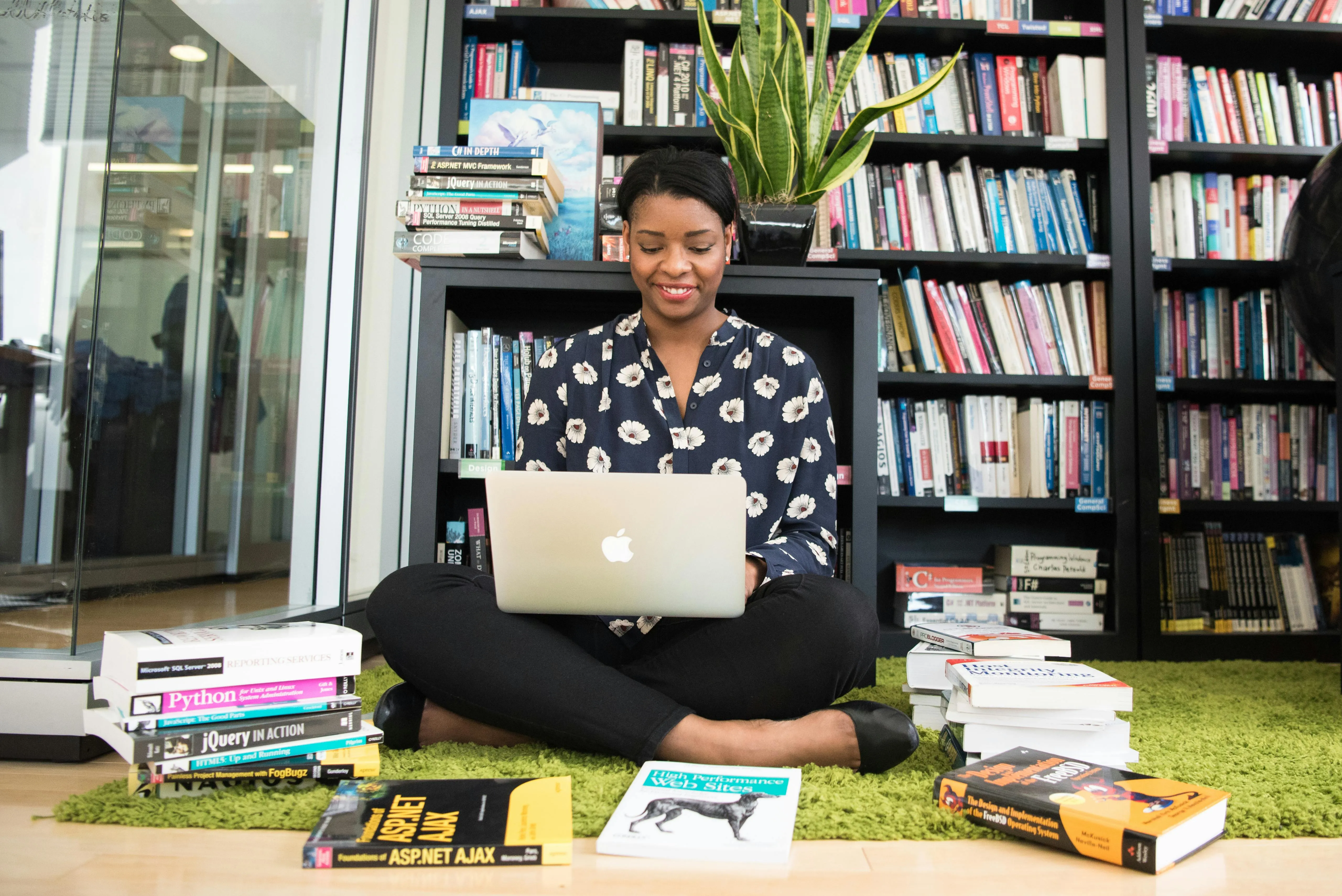 A woman sitting on the floor with her laptop and with books around her and a library shelf behind.