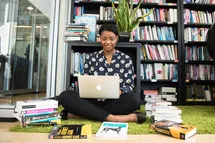 A woman sitting on the floor with her laptop and with books around her and a library shelf behind.