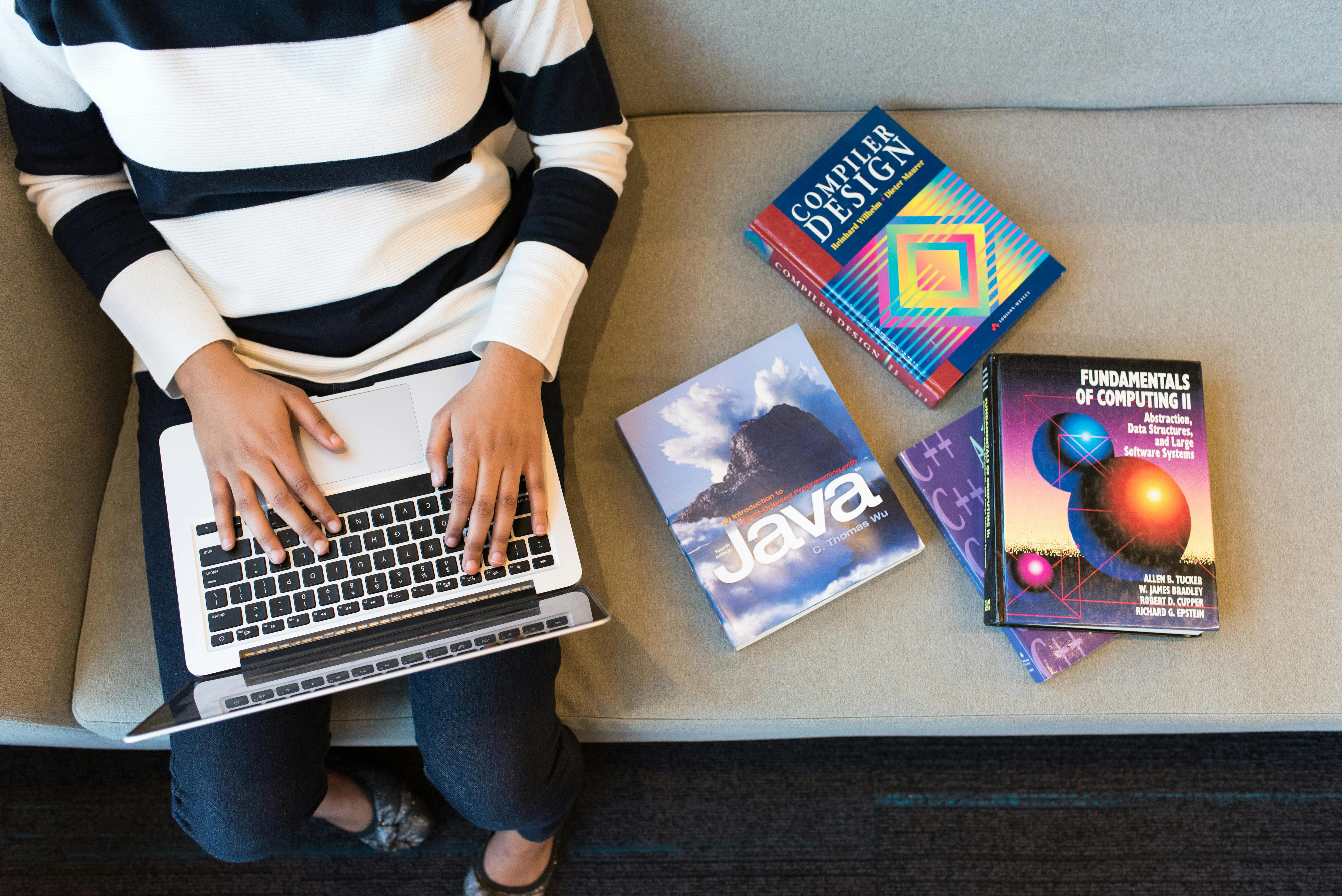 A person with a laptop siting on a sofa with java and design books on his left side, the image taken from above the person.