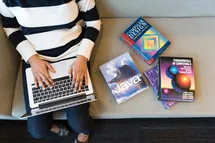 A person with a laptop siting on a sofa with java and design books on his left side, the image taken from above the person.