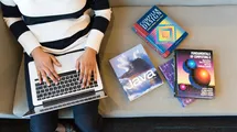 A person with a laptop siting on a sofa with java and design books on his left side, the image taken from above the person.
