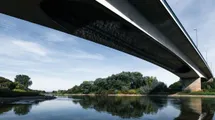 A photo taken from the riverbank looking up at the underside of a long concrete bridge spanning a calm river, with trees and sky reflected in the water below.
