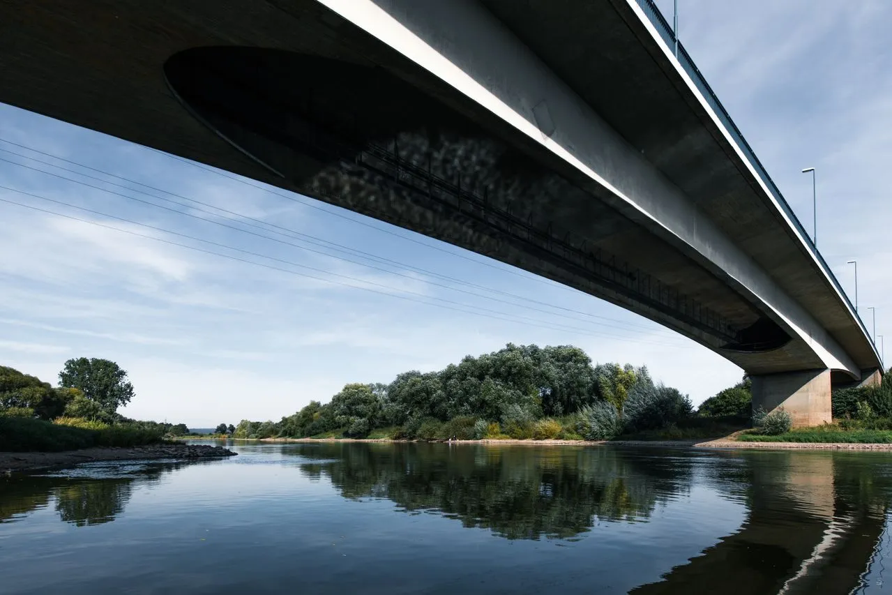 A photo taken from the riverbank looking up at the underside of a long concrete bridge spanning a calm river, with trees and sky reflected in the water below.